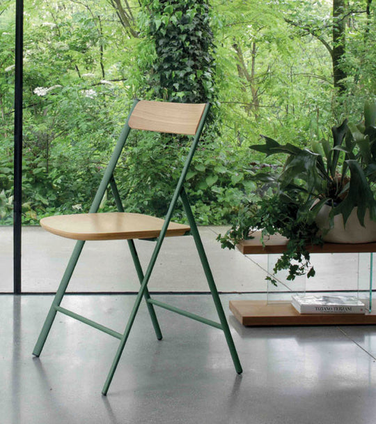 Joe Wood Foldable chair with wooden seat and green metal frame in a room with plants and a glass wall.