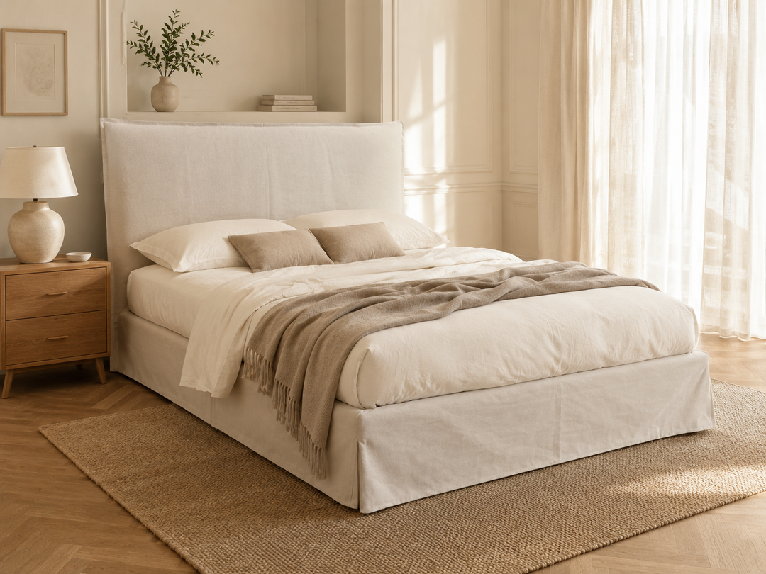 Shabby chic a storage bed dressed in cream bedding and a taupe throw blanket, set in a softly lit bedroom with its tall upholstered headboard, wooden nightstand, lamp, rug, and sheer curtains.