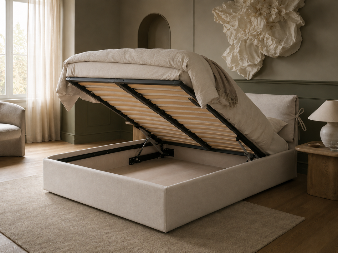 Cloude a light beige upholstered storage bed with its lift-up mattress platform open, showing the support slats and storage compartment, set in a styled bedroom with a side table, lamp, and neutral wall decor.