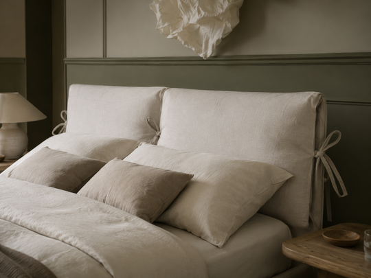 Cloude close-up of a headboard with a warm off-white fabric, styled with matching pillows and bedding in a softly lit bedroom.