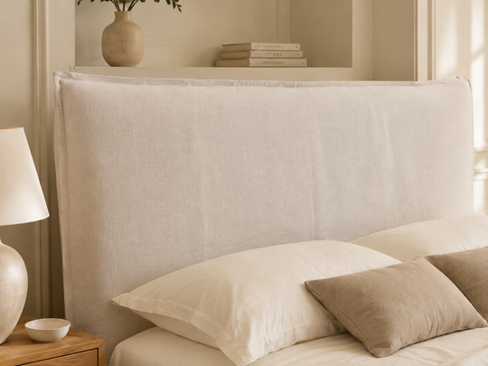 Shabby chic upholstered headboard in a warm off-white fabric, styled on a bed with cream pillows and a taupe accent pillow in a softly lit bedroom.