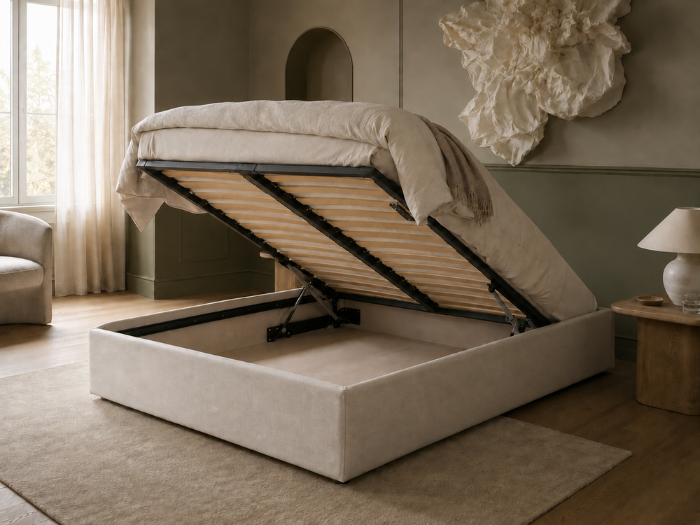 Ted a upholstered storage bed in a warm beige tone, shown with the lift-up mattress platform open to reveal the wooden slats and gas lift mechanism. The bed is styled in a modern, neutral-toned bedroom with a matching rug, side table, and lamp.