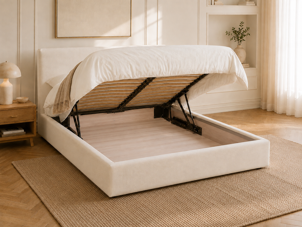 Teddy a low-profile upholstered storage ottoman bed in a warm cream or off-white fabric, shown with the mattress lifted to reveal the storage aread under the it. The bed is styled in a bright, modern bedroom with a woven rug, wooden floor, and a bedside table.